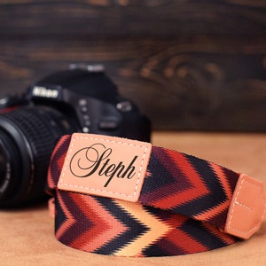 Può includere: Un cinturino per fotocamera con un motivo geometrico in tonalità di rosso, nero e arancione. Il cinturino ha un patch in pelle con il nome "Steph" stampato su di esso.