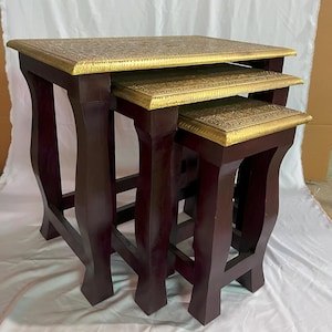May include: Set of three nesting tables. The tables have dark brown wooden legs and tops with a gold-colored, ornate design. The tables are of varying sizes, designed to fit one under the other. The tables are a decorative furniture item.