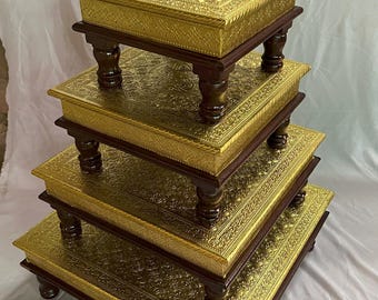 Può includere: Un set di quattro supporti decorativi in legno. Ogni supporto ha una superficie dorata, ornata e in rilievo. I supporti rettangolari hanno gambe e finiture marrone scuro. I supporti sono impilati, diminuendo di dimensioni.