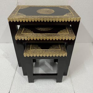 Puede incluir: Juego de tres mesas nido. Las mesas tienen un acabado marrón oscuro con adornos y detalles de metal dorado. Las tapas son cuadradas con un diseño circular central. Las mesas son de diferentes tamaños, diseñadas para encajar entre sí.