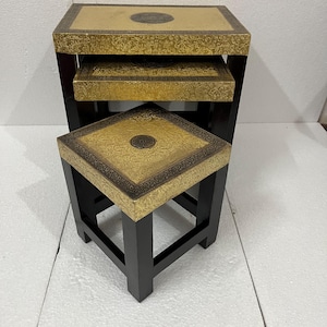 May include: Set of three nesting tables. Each table has a black frame and a gold-colored top with a decorative pattern. The tables are square and rectangular, designed to fit inside each other for storage.