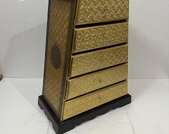 May include: A gold-colored chest of drawers with five drawers. The chest has a decorative floral pattern and is set on a black base. The sides of the chest are angled, and the top has a slight overhang.
