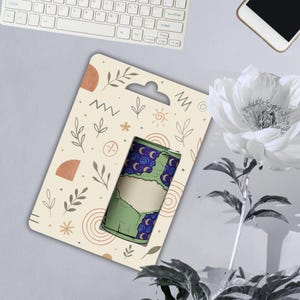 May include: A green and blue cylindrical product with a blank label, displayed on a patterned card with a hanging hole. The card has a beige background with abstract shapes and botanical illustrations. A white keyboard, phone, and flower are in the background.
