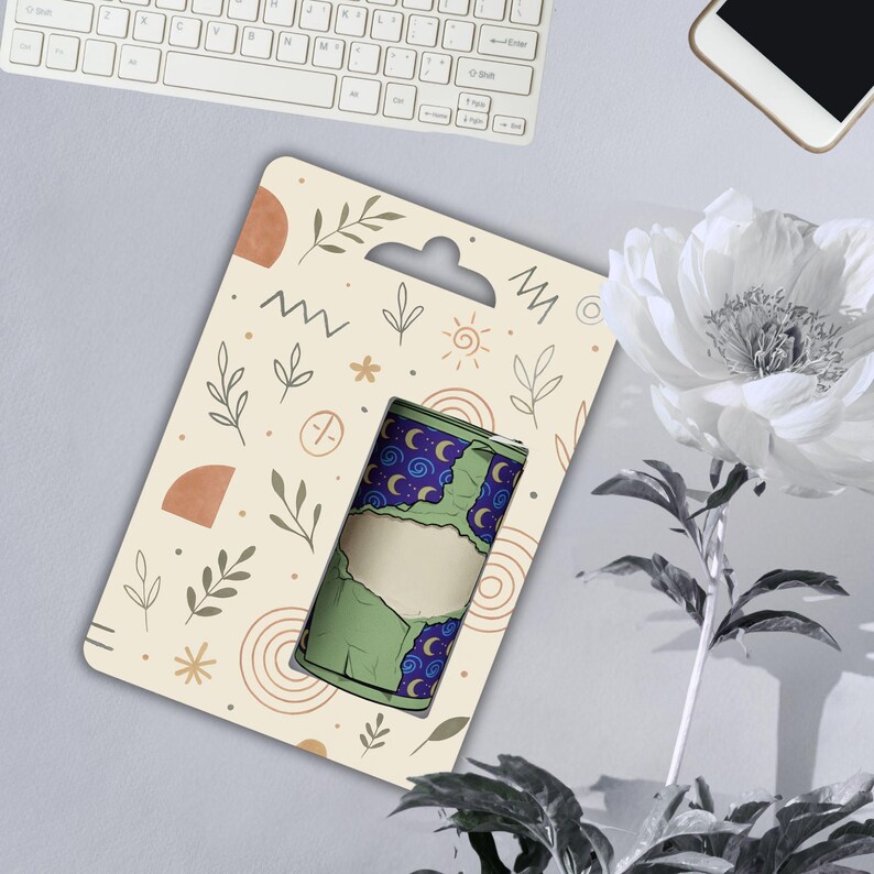 May include: A green and blue cylindrical product with a blank label, displayed on a patterned card with a hanging hole. The card has a beige background with abstract shapes and botanical illustrations. A white keyboard, phone, and flower are in the background.