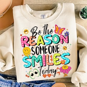 Könnte beinhalten: Ein cremefarbener Pullover mit einer bunten Grafik, auf der "Sei der Grund, warum jemand heute lächelt" steht. Die Grafik zeigt ein lächelndes Gesicht, einen Schmetterling, Blumen und andere fantasievolle Designs.