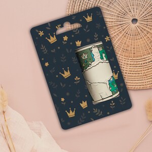 May include: A roll of decorative tape with a torn paper effect, featuring a green and blue pattern with crowns and a blank label. The tape is displayed on a navy blue card with gold crowns and floral accents.