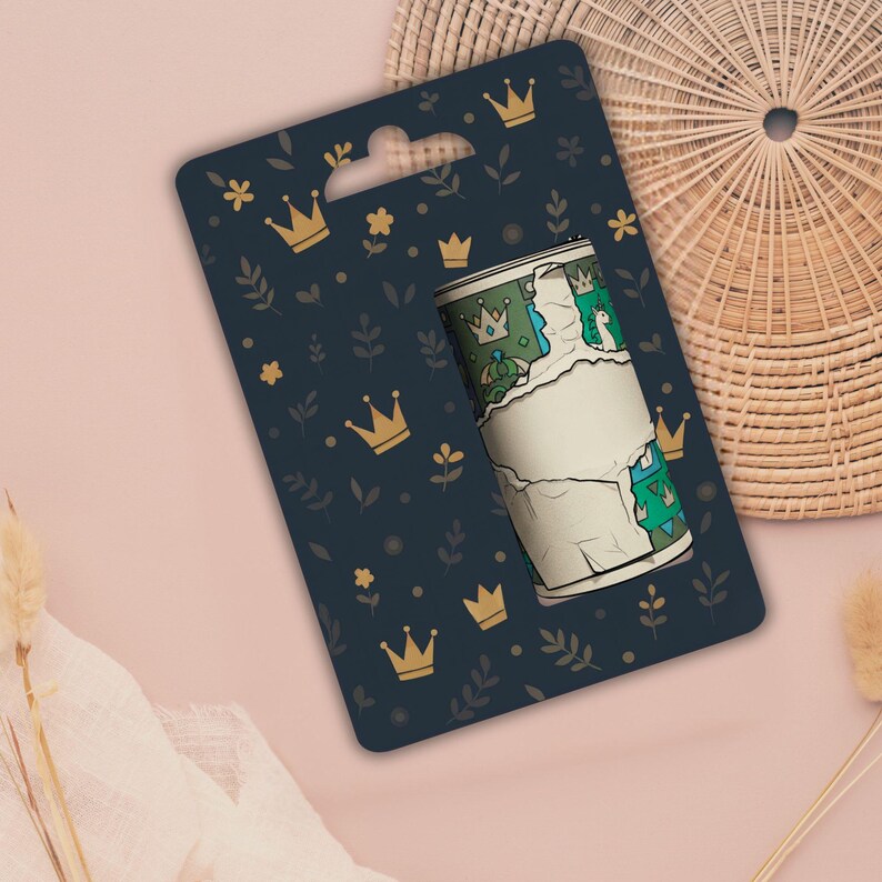May include: A roll of decorative tape with a torn paper effect, featuring a green and blue pattern with crowns and a blank label. The tape is displayed on a navy blue card with gold crowns and floral accents.