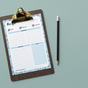 Puede incluir: Una plantilla de receta azul y blanca en un portapapeles marr&oacute;n con un clip dorado. La plantilla tiene secciones para porciones, tiempo de preparaci&oacute;n, tiempo de cocci&oacute;n, ingredientes, instrucciones y notas. Un l&aacute;piz negro est&aacute; acostado junto al portapapeles.