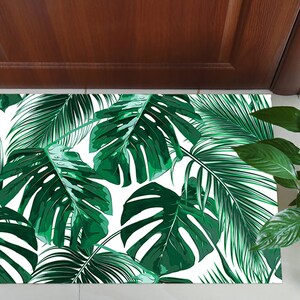 May include: A rectangular doormat featuring a vibrant tropical leaf pattern in shades of green and white. The design includes large monstera leaves and other tropical foliage. The mat is placed in front of a wooden door and next to a potted plant.