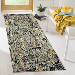 Puede incluir: Una alfombra rectangular con un diseño abstracto. La alfombra presenta un fondo gris con salpicaduras de pintura negra, amarilla, roja y azul. La alfombra está colocada sobre un suelo de madera blanco, cerca de una puerta y un banco.