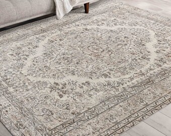 May include: A rectangular rug featuring a detailed floral design in beige, grey, and brown tones. The rug has a central medallion and a border with geometric patterns. It is placed on a light-coloured wooden floor.