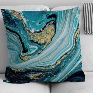 May include: Square throw pillow with an abstract design in shades of teal, blue, black, and white, accented with gold metallic swirls. The pillow is placed on a white sofa, showcasing its decorative pattern.
