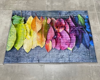 May include: A colorful doormat with a design of autumn leaves in shades of yellow, orange, red, purple, and blue. The leaves are arranged in a row across the doormat, which has a dark gray background.