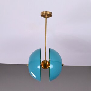 Op de afbeelding: Een moderne hanglamp met twee tealblauwe halfronde lampenkappen die met een gouden bol zijn verbonden. De lamp wordt aan het plafond gehangen met een gouden stang.