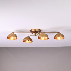 Könnte beinhalten: Eine Deckenleuchte in Messingoptik mit vier halbkugelförmigen Schirmen, die jeweils eine warmtonige Glühbirne enthalten. Die Leuchte ist an einer weißen Decke befestigt. Das Design ist modern und minimalistisch. Die Breite beträgt ca. 60 cm.
