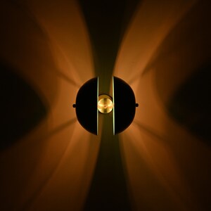 Op de afbeelding: Een zwarte en gouden wandlamp met een rond, gesplitst ontwerp. De lamp geeft een warm licht aan de muur.