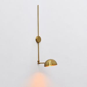 Könnte beinhalten: Eine Wandleuchte aus Messing mit einer langen vertikalen Stange, einer runden Montageplatte und einem verstellbaren Arm, der zu einem kuppelförmigen Schirm führt. Die Lampe strahlt ein warmes, sanftes Licht von der Unterseite des goldenen Schirms aus.