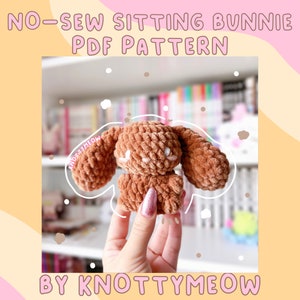 Peut inclure: Un lapin crocheté brun assis avec des yeux blancs. Le lapin est tenu par une main. Le texte de l'image est "NO-SEW SITTING BUNNIE PDF PATTERN BY KNOTTYMEOW".