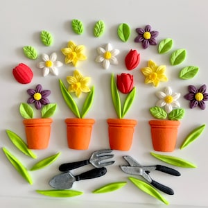May include: A set of edible cake decorations featuring three potted plants with flowers, two gardening tools, and several leaves. The flowers are yellow daffodils, white daisies, red tulips, and purple pansies. The tools are a trowel and a pair of pruning shears.