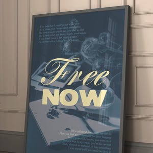 May include: Framed art print with the words "Free Now" in gold lettering. The print has a dark blue background with handwritten text and a pen and notebook visible. The frame is gray.