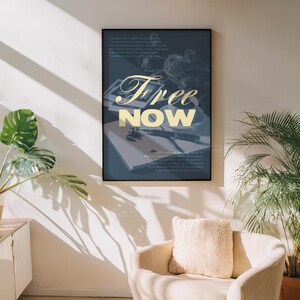 May include: A framed art print with the words "Free Now" in gold script. The artwork features a dark blue background with a notebook, pen, and other objects. The print is displayed on a white wall above a beige armchair.