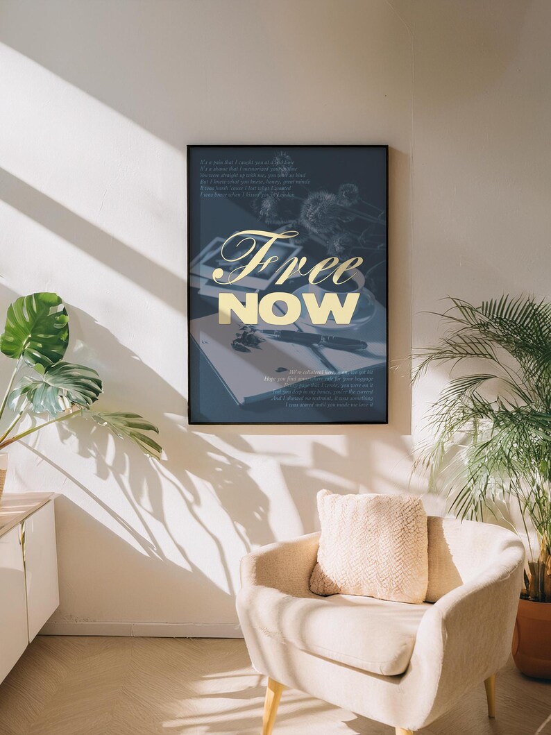 May include: A framed art print with the words "Free Now" in gold script. The artwork features a dark blue background with a notebook, pen, and other objects. The print is displayed on a white wall above a beige armchair.