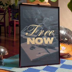 May include: Framed artwork with the words "Free NOW" in gold lettering. The artwork has a dark background with handwritten text and a drawing. The frame is black, and the artwork is displayed on a blue and green patterned rug.
