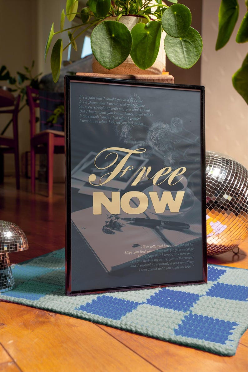 May include: Framed artwork with the words "Free NOW" in gold lettering. The artwork has a dark background with handwritten text and a drawing. The frame is black, and the artwork is displayed on a blue and green patterned rug.