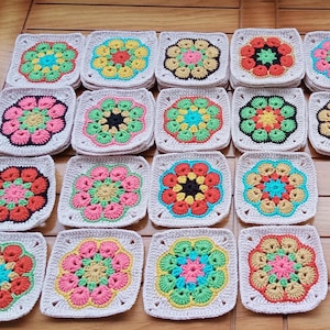 May include: A collection of sixteen square crochet pieces with floral designs. Each square features a central flower in various colors like pink, orange, yellow, and green, surrounded by a cream-colored border. The squares are arranged on a wooden surface.
