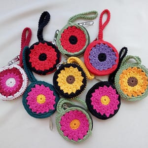 Puede incluir: Una colección de estuches redondos de ganchillo con cremallera y correas de muñeca, cada uno con un patrón floral multicolor. Los estuches muestran varias combinaciones de colores que incluyen hilo rojo, amarillo, rosa, morado, naranja, verde, negro y blanco.