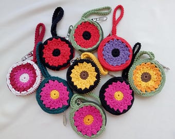 May include: A collection of round, crocheted zip pouches with wrist straps, each featuring a multi-coloured floral pattern. The pouches display various colour combinations including red, yellow, pink, purple, orange, green, black, and white yarn.