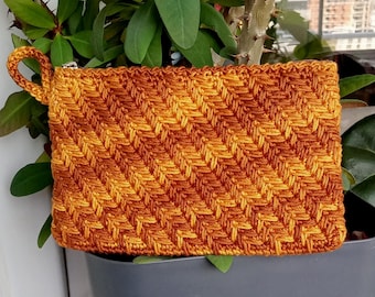 Puede incluir: Un neceser rectangular de ganchillo hecho a mano en tonos naranja y marrón. El neceser presenta un patrón de tejido diagonal texturizado y un pequeño asa de bucle. Una cremallera plateada asegura la abertura superior. Se muestra frente a follaje verde.