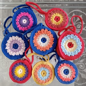 Puede incluir: Una colección de ocho monederos redondos de ganchillo con cremallera y vibrantes patrones florales multicolores. Cada monedero incluye una correa de muñeca de ganchillo a juego y un cierre de cremallera metálico plateado en varios colores.