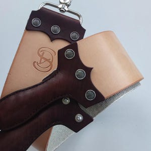 Może przedstawiać: Skórzany pas do ostrzenia z ciemnobrązowym uchwytem i jasnobrązową skórzaną powierzchnią. Pas ma srebrną klamrę i nity. Skórzana powierzchnia ma wytłoczony wzór. Ten przedmiot służy do ostrzenia brzytew.