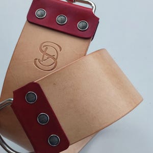 May include: A tan leather strop with a red leather end and a silver hook. The strop has three silver rivets on each end and a silver logo in the center.