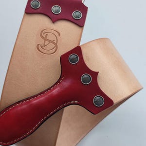 Op de afbeelding: Een rode en bruine leren strop met een zilveren haak en klinknagels. De strop heeft een rode leren handgreep en is ontworpen om scheermessen te slijpen.