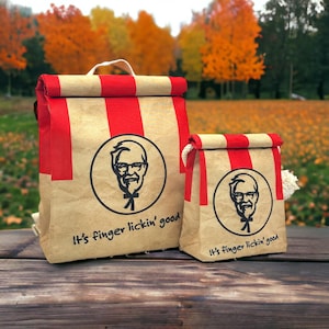 Puede incluir: Dos bolsas de almuerzo de color beige con rayas verticales rojas y cierre enrollable. Cada bolsa presenta un logotipo circular negro del Coronel Sanders y el texto "It's finger lickin' good" impreso en negro en la parte frontal.