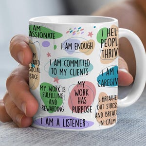 Könnte beinhalten: Weiße Keramiktasse mit bunten abstrakten Formen und positiven Affirmationen in schwarzem Text. Der Text beinhaltet "Ich bin genug", "Ich bin meinen Kunden verpflichtet", "Meine Arbeit hat einen Sinn", und "Ich bin ein Zuhörer".