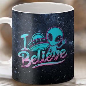 Pode incluir: Caneca de cerâmica branca com fundo preto que apresenta um alienígena azul com um disco voador e o texto "I Believe" em letras rosa e azul.