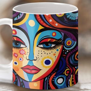 May include: White ceramic mug with a colorful abstract design of a woman's face. The design features bright colors like blue, red, yellow, and green.