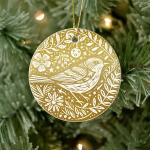 May include: A round, gold-colored Christmas ornament with a white bird and floral design. The bird is surrounded by leaves, flowers, and stars. A gold string is attached for hanging. The ornament is displayed on a Christmas tree.