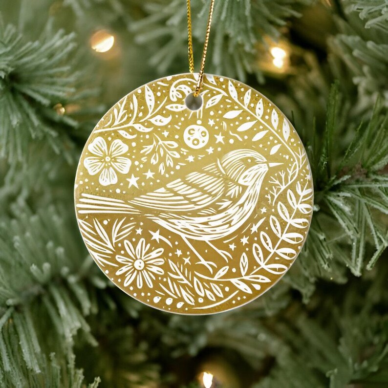 May include: A round, gold-colored Christmas ornament with a white bird and floral design. The bird is surrounded by leaves, flowers, and stars. A gold string is attached for hanging. The ornament is displayed on a Christmas tree.