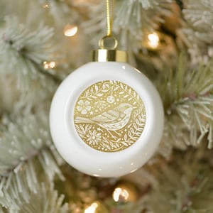 May include: White ceramic Christmas ornament with a gold bird and floral design. The ornament has a gold cap and a gold ribbon for hanging. The ornament is hanging on a Christmas tree with white and green branches.