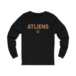 May include: Black long-sleeve shirt with the word "ATLIENS" in a tan, textured font across the chest. Below the text is a small circular graphic with the letters "ATL" inside. The shirt is made of a soft material.