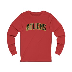 May include: Red long-sleeve t-shirt with the word "ATLIENS" printed in black with a yellow outline.