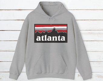 Puede incluir: Una sudadera con capucha gris con un gráfico rectangular negro que presenta una cordillera estilizada y la palabra "atlanta" en blanco.