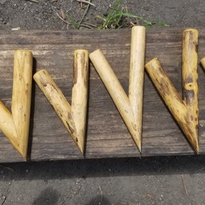 Könnte beinhalten: Acht V-förmige Holzpfähle, jeder mit einer spitzen Spitze, sind in einer Reihe auf einer Holzoberfläche angeordnet. Die Pfähle sind hellbraun und haben eine natürliche, rustikale Oberfläche.