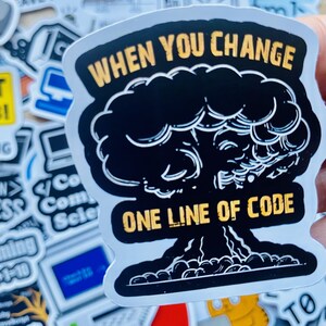 Może przedstawiać: Biała naklejka z czarno-białym rysunkiem eksplodującego grzyba atomowego. Naklejka zawiera napis "WHEN YOU CHANGE ONE LINE OF CODE" w kolorze ż&oacute;łtym.