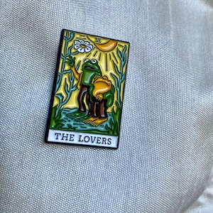 May include: A green and yellow enamel pin depicting the Lovers tarot card with two frogs, one holding a flower, and the text "THE LOVERS".