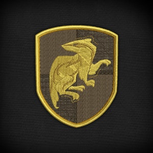 Könnte beinhalten: Ein gold gestickter Aufnäher mit braunem Hintergrund. Der Aufnäher zeigt ein stilisiertes Bild einer mythischen Kreatur, möglicherweise ein Greif, mit langem Hals und Flügeln.